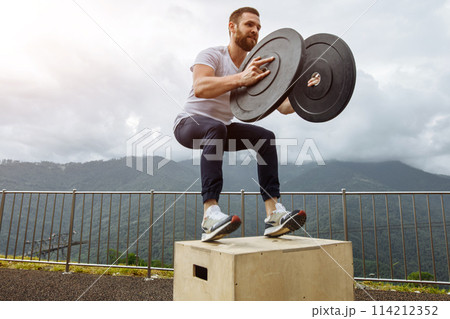 Strong male athlete doing box jumps with two plates outdoor on top of mountain. 114212352