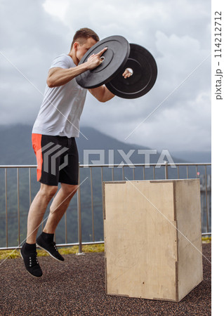 Strong male athlete doing box jumps with two plates outdoor on top of mountain. Strong male athlete doing box jumps with two plates outdoor on top of mountain. 114212712