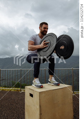 Strong male athlete doing box jumps with two plates outdoor on top of mountain. 114212714