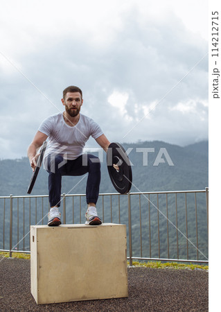 Strong male athlete doing box jumps with two plates outdoor on top of mountain. 114212715