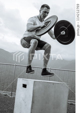 Strong male athlete doing box jumps with two plates outdoor on top of mountain. 114212769