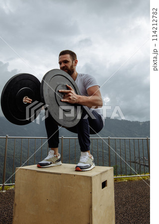 Strong male athlete doing box jumps with two plates outdoor on top of mountain. 114212782
