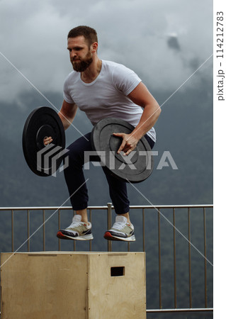Strong male athlete doing box jumps with two plates outdoor on top of mountain. Strong male athlete doing box jumps with two plates outdoor on top of mountain. 114212783