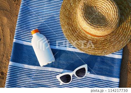 Beach scene with sunblock bottle, striped towel, stylish sunglasses, straw hat on sandy background. Accessories for skin protection against summer sun, holiday vacation concept by sea. Beach scene with sunblock bottle, striped towel, stylish sunglasses, straw hat on sandy background. Accessories for skin protection against summer sun, holiday vacation concept by sea. 114217730
