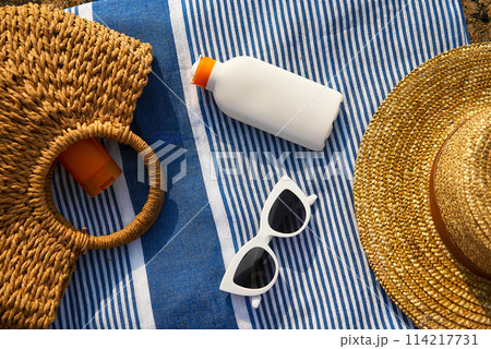 Overhead shot of summer essentials on striped towel with white sunscreen bottle, woven straw hats, stylish white sunglasses, ready for sun-safe beach day by the ocean. 114217731