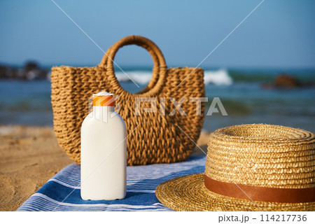 Wicker bag, sun hat, sunscreen bottle on striped towel at sunny seaside. Summer accessories prepare for sunbath, promote skin safety, holiday essentials, beach day gear with ocean backdrop. 114217736
