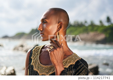 Outrageous black lgbtq person in luxury jewelry poses on scenic ocean beach. Non-binary ethnic fashion model in posh outfit stands gracefully at picturesque location near sea, lighthouse. Pride month. Outrageous black lgbtq person in luxury jewelry poses on scenic ocean beach. Non-binary ethnic fashion model in posh outfit stands gracefully at picturesque location near sea, lighthouse. Pride month. 114217741