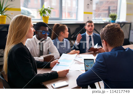 Multiracial business team in office analyze data on charts, diagrams. Caucasian woman, African man, and colleagues plan strategies. Corporate group discuss growth, sales, marketing ideas in meeting. 114217749