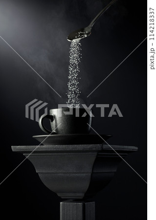 Sugar is poured into a black cup of coffee on a black background. 114218337