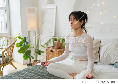 Yoga mindfulness meditation. Young healthy woman practicing yoga in bedroom at home. Woman sitting in lotus pose on bed meditating smiling relaxing indoor. Girl doing breathing practice. Yoga at home Yoga mindfulness meditation. Young healthy woman practicing yoga in bedroom at home. Woman sitting in lotus pose on bed meditating smiling relaxing indoor. Girl doing breathing practice. Yoga at home 114219106