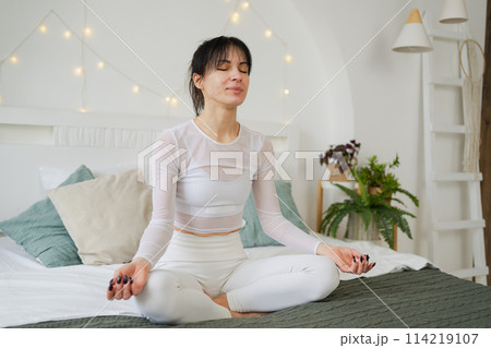Yoga mindfulness meditation. Young healthy woman practicing yoga in bedroom at home. Woman sitting in lotus pose on bed meditating smiling relaxing indoor. Girl doing breathing practice. Yoga at home 114219107
