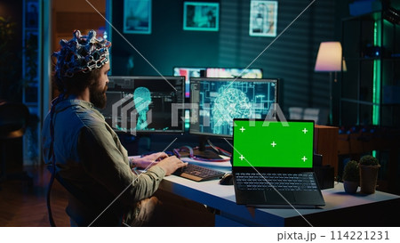 Engineer with EEG headset on programming brain transfer into computer virtual world next to green screen laptop. Transhumanist using neuroscience to gain digital soul, mockup device, camera B Engineer with EEG headset on programming brain transfer into computer virtual world next to green screen laptop. Transhumanist using neuroscience to gain digital soul, mockup device, camera B 114221231
