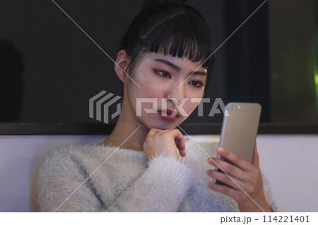 夜自宅でスマートフォンの画面を見て考え込む若い女性 夜自宅でスマートフォンの画面を見て考え込む若い女性 114221401