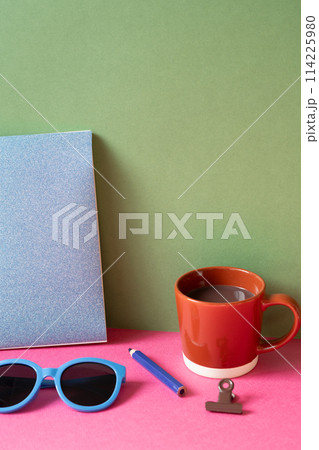 Desktop workspace. notebook, eyeglasses, colored pencil, clip, cup of hot chocolate on pink desk. khaki green wall background Desktop workspace. notebook, eyeglasses, colored pencil, clip, cup of hot chocolate on pink desk. khaki green wall background 114225980