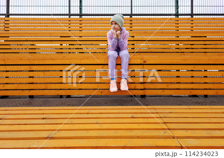 A girl sits on a bench in a sports tribune with her hands on her knees. 114234023