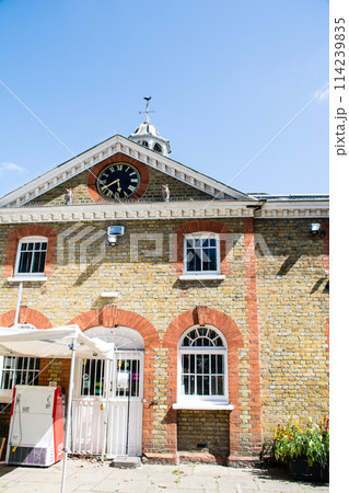 明るい日差しの下の大時計の付いた歴史的なレンガ造りの建物 ロンドンの西のラベンズコート公園にて 明るい日差しの下の大時計の付いた歴史的なレンガ造りの建物 ロンドンの西のラベンズコート公園にて 114239835