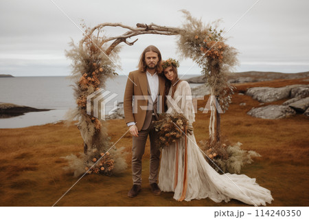 Boho style bride and groom are holding hands in the background of romantic wedding arch at the sea bay and mountains. 114240350