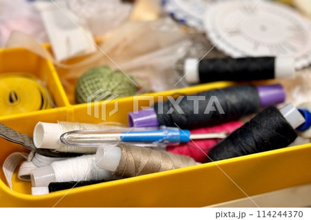 A lot of sewing items such as buttons, colorful fabrics, scissors, measuring tape, thimble, spools of thread on sewing pattern located in plastic box on the sofa. Close-up 114244370