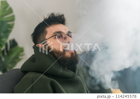 Portrait bearded man is smoking hookah at home and blowing cloud of smoke, copy space and place for text - chill time and resting concept Portrait bearded man is smoking hookah at home and blowing cloud of smoke, copy space and place for text - chill time and resting concept 114245578