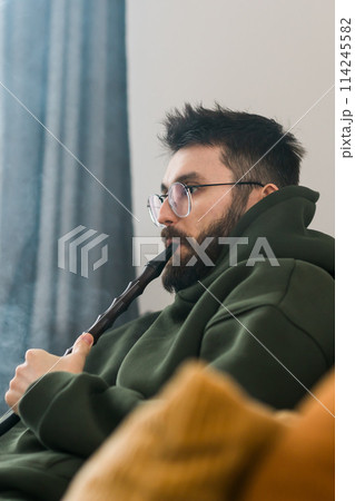 Portrait bearded man is smoking hookah at home and blowing cloud of smoke, copy space and place for text - chill time and resting concept 114245582