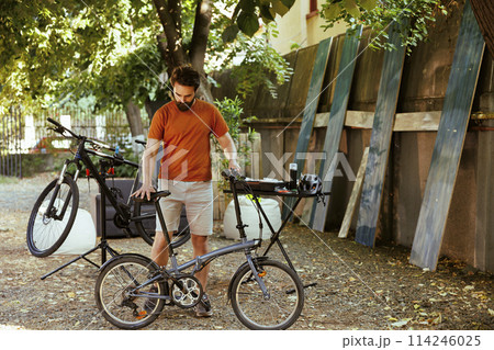 Caucasian male cyclist doing home maintenance in his yard surrounded by collection of professional cycling gear. Sports-loving man ensuring safety of bicycle after repairing. Caucasian male cyclist doing home maintenance in his yard surrounded by collection of professional cycling gear. Sports-loving man ensuring safety of bicycle after repairing. 114246025
