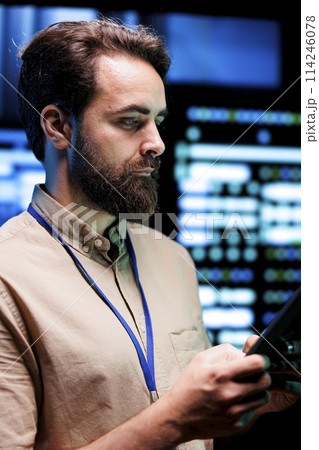 Programmer doing thorough system examination, checking data on tablet of needed debugging in server mainframes housing advanced data storage hardware supporting complex computer operations 114246078