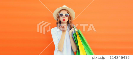 Stylish beautiful young woman with shopping bags blowing a kiss in summer straw hat on orange 114246453