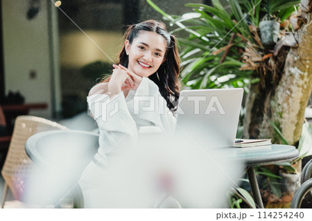 Happy young woman enjoying work on her laptop at a cozy outdoor cafe surrounded by plants. 114254240