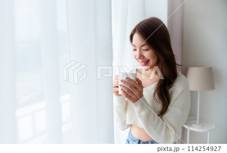 Young asian woman standing beside window and holding mug in bedroom at home Young asian woman standing beside window and holding mug in bedroom at home 114254927