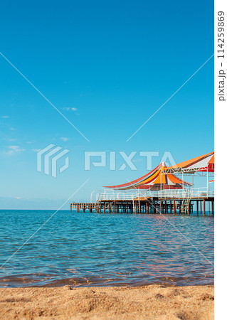 Wooden pier with orange-red awning over water of sea, lake, ocean with beach without people. Travel, vacation, summer background. Vertical. Copy space 114259869