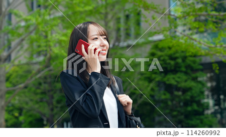新緑の街で歩きながらスマホで通話する女性ビジネスパーソン 114260992
