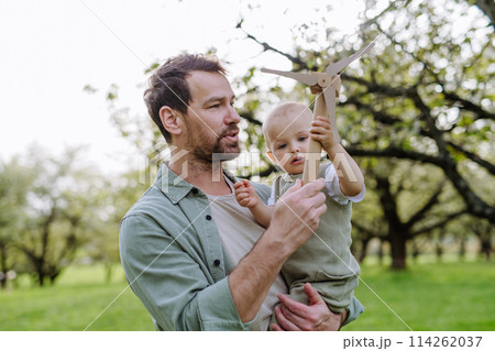 Baby and father holding wind turbine model, outdoors in park. Concept of renewable wind energy, sustainable future for the next generation. 114262037