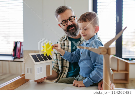 Father explaining renewable green energy, teaching son about sustainable lifestyle. Playing with model of house with solar panels, wind turibine at home. Learning through play. 114262485