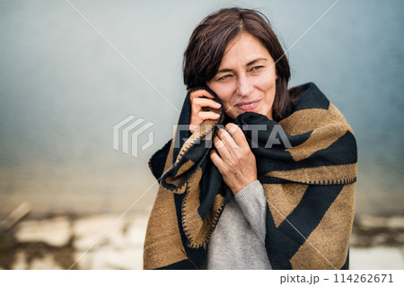 Portrait of beautiful mature woman with blanket around shoulders, standing by lake. 114262671