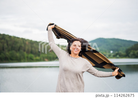 Portrait of beautiful mature woman holding blanket in air, wind blowing in hair, standing by lake. 114262675
