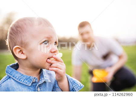 Little boy eating snack outdoors. Father waiting for him, sitting in grass. Father's day concept. Family time outdoors during warm spring day. Little boy eating snack outdoors. Father waiting for him, sitting in grass. Father's day concept. Family time outdoors during warm spring day. 114262960
