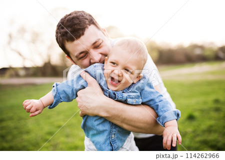 Father holding little toddler boy kissing him, making him laugh, having fun during warm spring day. Father's day concept. Father holding little toddler boy kissing him, making him laugh, having fun during warm spring day. Father's day concept. 114262966