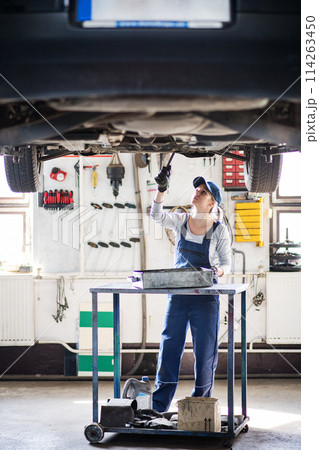 Female auto mechanic elevating car on car lift, changing oil. Beautiful woman working in a garage, wearing blue coveralls. Female auto mechanic elevating car on car lift, changing oil. Beautiful woman working in a garage, wearing blue coveralls. 114263450
