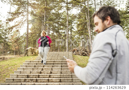 Overweight woman running down the stairs, personal trainer checking her performance. Exercising outdoors for people with obesity, support from friend, fitness coach. 114263629
