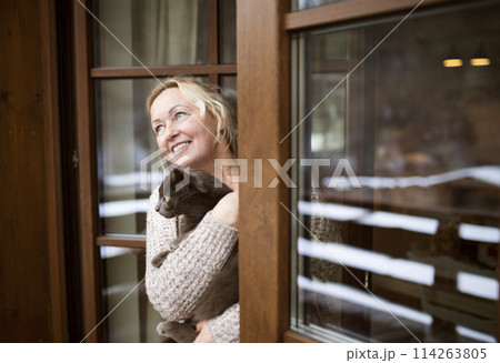 Beautiful mature woman at home, holding, petting her cat, looking from window. Older woman living alone, enjoying peaceful weekend day 114263805