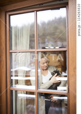 Beautiful mature woman at home, holding, petting her cat, looking through the window. Older woman living alone, enjoying peaceful weekend day Beautiful mature woman at home, holding, petting her cat, looking through the window. Older woman living alone, enjoying peaceful weekend day 114263810