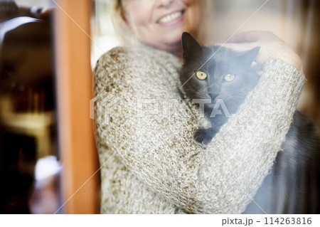 Beautiful mature woman at home, holding, petting her cat, looking through the window. Older woman living alone, enjoying peaceful weekend day Beautiful mature woman at home, holding, petting her cat, looking through the window. Older woman living alone, enjoying peaceful weekend day 114263816