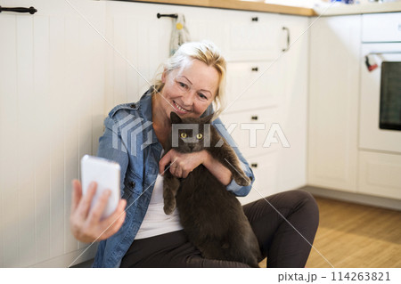 Beautiful mature woman at home, taking selfie with her cat. Older woman living alone, enjoying peaceful weekend day Beautiful mature woman at home, taking selfie with her cat. Older woman living alone, enjoying peaceful weekend day 114263821