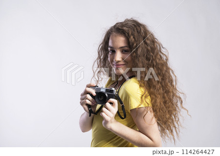 Portrait of a gorgeous teenage girl with curly hair holding camera. Studio shot, white background with copy space 114264427