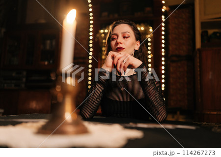 Low-angle portrait of female soothsayer dealing Tarot cards forecasting future for client by light of burning candles. Fortune-telling having divination session with tarot cards at dark esoteric room. Low-angle portrait of female soothsayer dealing Tarot cards forecasting future for client by light of burning candles. Fortune-telling having divination session with tarot cards at dark esoteric room. 114267273