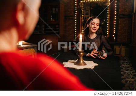 View from shoulder of enigmatic fortune teller reading tarot cards to middle-aged woman client during spiritual ceremony in dark room by light of burning candle. Concept of esoteric and predictions. 114267274