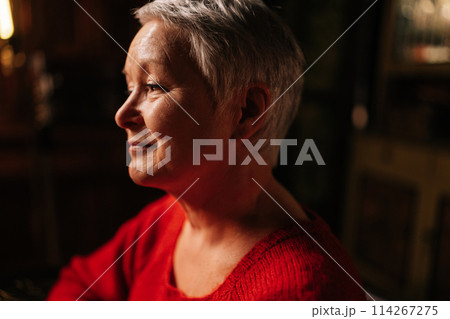 Close-up face of positive gray-haired senior adult woman in casual red sweater sitting at table in dark room by light of burning candle, smiling looking away. Close-up face of positive gray-haired senior adult woman in casual red sweater sitting at table in dark room by light of burning candle, smiling looking away. 114267275