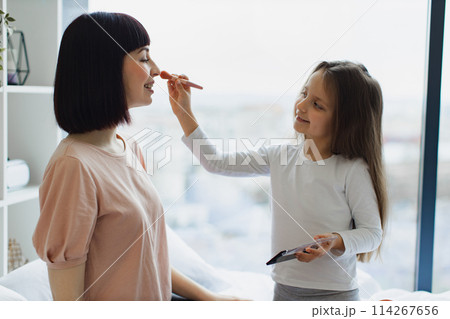 Close up portrait of Caucasian daughter applying powder on her young mom's face. Close up portrait of Caucasian daughter applying powder on her young mom's face. 114267656