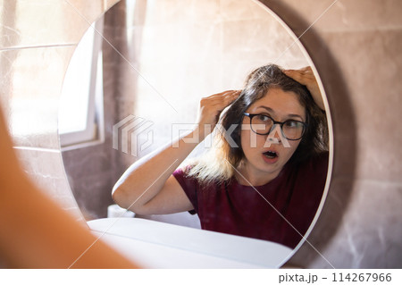 Gray haired surprised caucasian middle aged woman looking at grey hair head in mirror reflection. Early gray hair concept Gray haired surprised caucasian middle aged woman looking at grey hair head in mirror reflection. Early gray hair concept 114267966