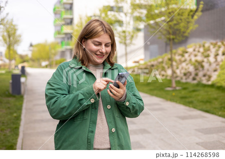 30s cheerful woman tourist dressed in casual look holding smartphone gadget in hand for communicate on the city street. Happy woman chatting on phone and relaxing outdoors. Urban, people concept. 30s cheerful woman tourist dressed in casual look holding smartphone gadget in hand for communicate on the city street. Happy woman chatting on phone and relaxing outdoors. Urban, people concept. 114268598